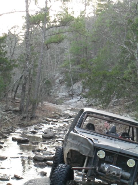 1986 Ford Bronco - A modified off-road vehicle navigates a shallow, rocky stream surrounded by trees and boulders.