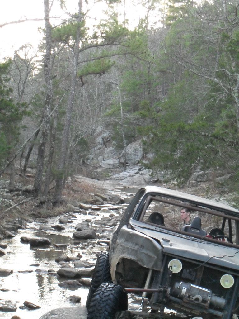 Off-road vehicle with roll cage driving through a rocky stream bed in a wooded area