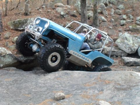 1986 Ford Bronco - A blue off-road jeep navigates a rocky terrain with two occupants inside.