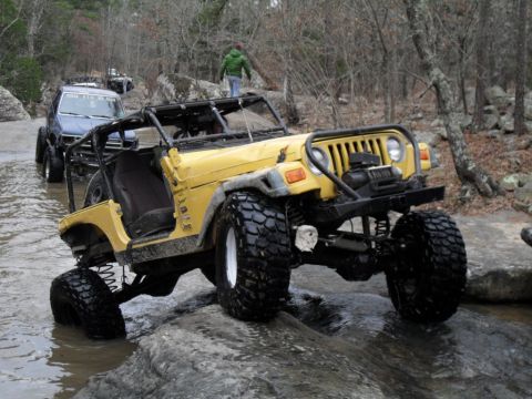 1986 Ford Bronco - A yellow Jeep Wrangler drives through a rocky stream with other vehicles following behind.