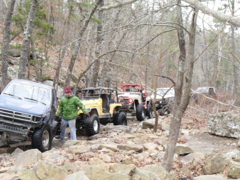 1986 Ford Bronco - Multiple off-road vehicles traverse a challenging rocky path through a wooded area.
