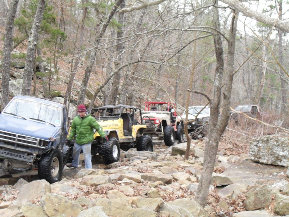 Several off-road vehicles navigating a rocky, wooded trail. A blue Toyota truck is in the foreground