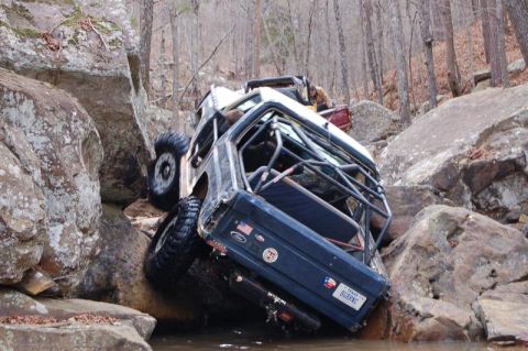 1986 Ford Bronco - This modified off-road vehicle is wedged between large rocks in a wooded area, with another vehicle visible behind it.