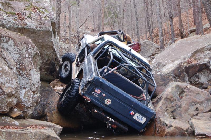 Off-road vehicle tilted on rocks, stuck in a stream. Another vehicle is behind it
