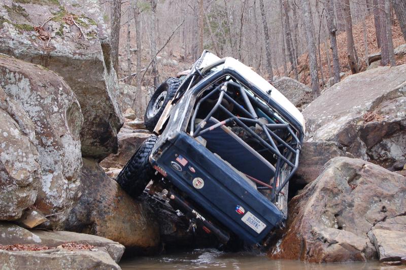 Blue off-road vehicle with a roll cage tilted on rocks in a stream