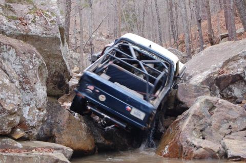 1986 Ford Bronco - Rear view of a dark blue full-size Bronco with a white roof and a black internal roll cage climbing nose-up out of a rocky creek bed, water…