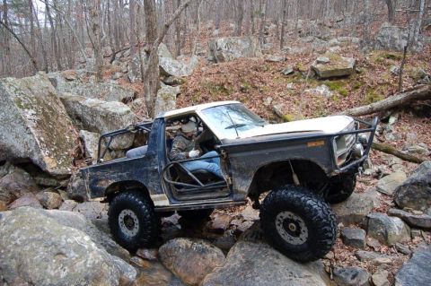 1986 Ford Bronco - High 3/4-front view of a doorless, topless dark blue and white 1980s Ford full-size 4x4 flexing over wet boulders on a rocky winter trail,…
