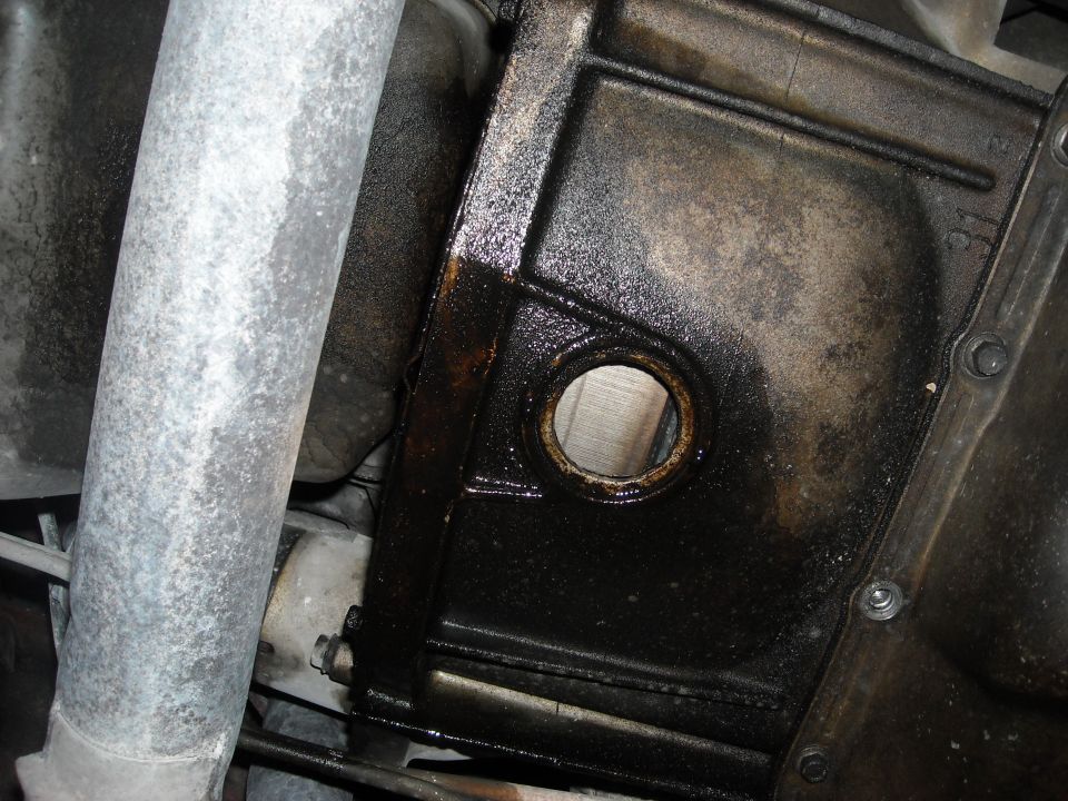 Underbody view of oily black pan with round inspection hole next to a silver exhaust pipe