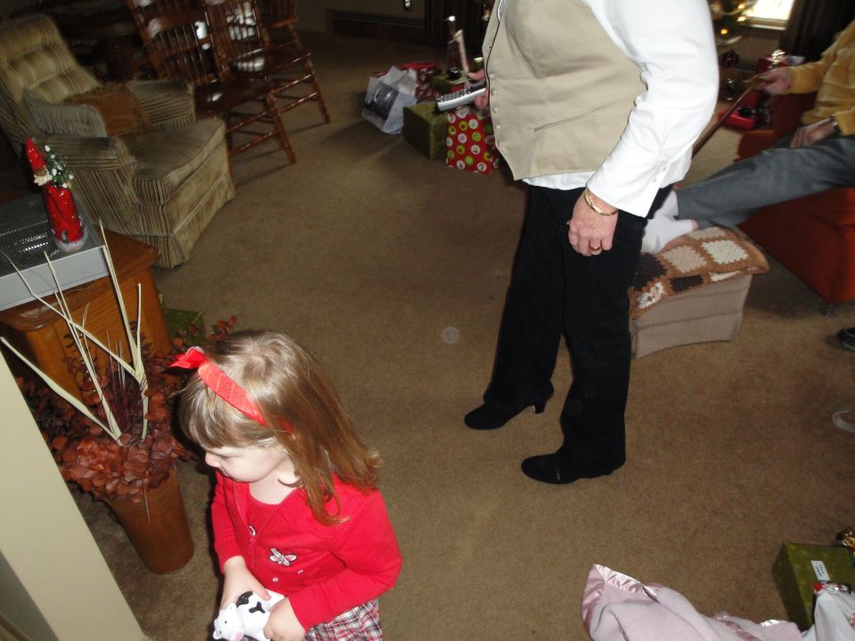 Child in red shirt looks down at toy, while adults stand nearby in a room with Christmas gifts