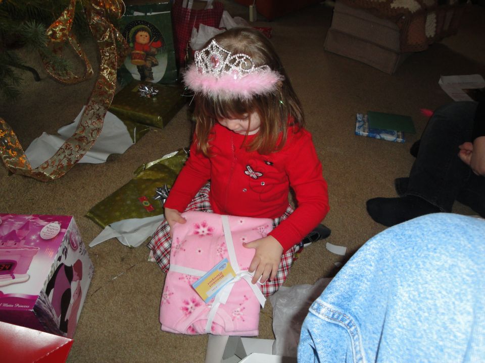 Young girl wearing a tiara opens a pink gift next to a Christmas tree with presents scattered around