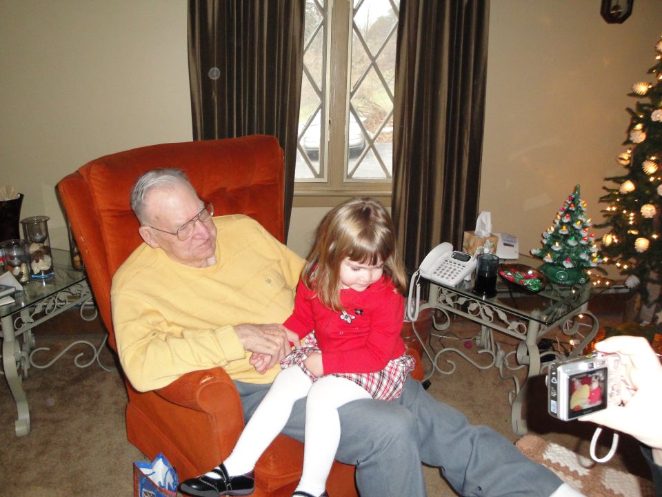 Man in yellow shirt and child in red sweater sit in an orange chair, with a Christmas tree in the background