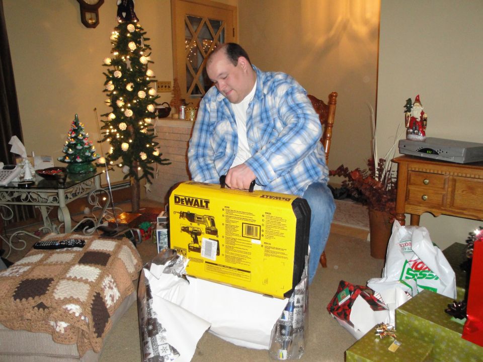 Man opening a yellow DeWalt drill set box, Christmas tree and presents in background