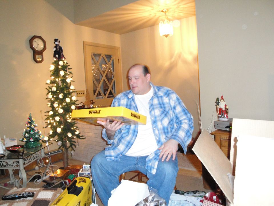 Man in blue plaid shirt holding yellow DeWalt box in a room with a Christmas tree