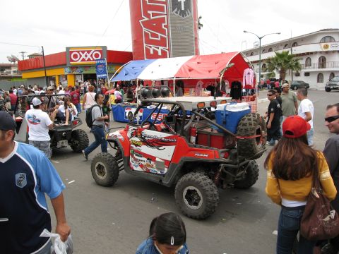 1986 Ford Bronco - An off-road buggy with racing decals is parked on a street during an event, with spectators and tents around.