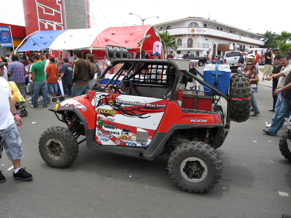 Side view of a red and white UTV with 'DragonFire Racing' graphics and sponsor decals. It has a roll cage, spare tire, and cooler rack