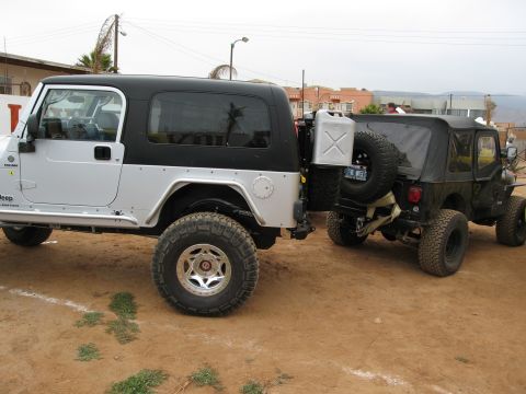 1986 Ford Bronco - Two Jeeps are parked on a dirt lot, one with a mounted gas can on the rear.