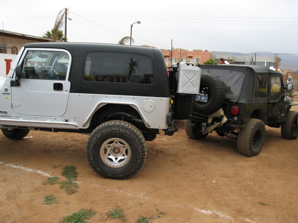 Two Jeeps, one silver and black, the other black, parked on dirt. The silver Jeep has a gas can mounted on the rear