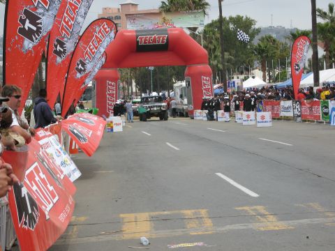 1986 Ford Bronco - A crowd gathers at the start of an off-road race event, marked by an inflatable arch and sponsor banners.