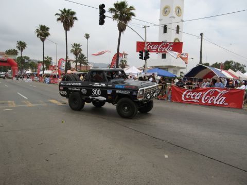 1986 Ford Bronco - A modified black Ford Bronco races past spectators and Coca-Cola banners under a cloudy sky.
