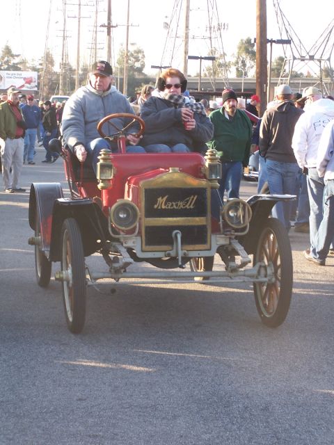 1990 Ford Bronco - A vintage Maxwell automobile is driven by two people, with onlookers in the background.