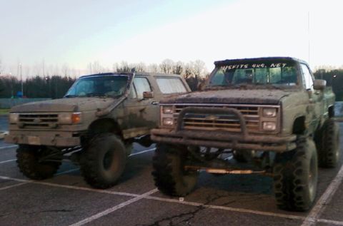 1990 Ford Bronco - Two heavily modified and mud-splattered trucks are parked in a lot, suggesting recent off-road activity.
