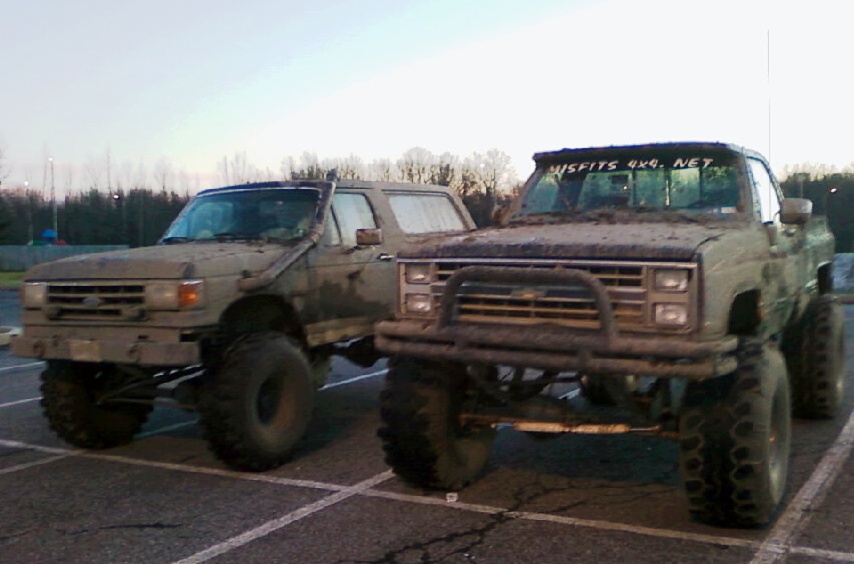 Two mud-covered trucks parked side-by-side, one is a Chevrolet pickup, the other appears to be an older Ford SUV