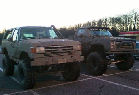 1990 Ford Bronco - A muddy 1990 Ford Bronco is parked next to a lifted pickup truck, both appearing ready for off-road adventures.