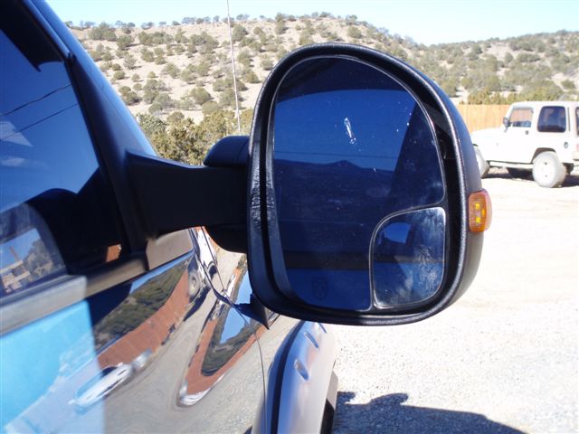 Truck's side mirror reflecting a hilly landscape. Another white SUV is parked nearby