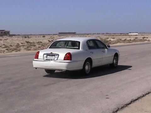 2002 Lincoln Town Car - A white sedan drives down a paved road in a dry, arid landscape under a clear blue sky.
