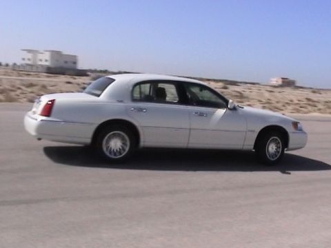 2002 Lincoln Town Car - A white sedan is captured in motion on a paved road with a desert landscape and buildings in the distance.