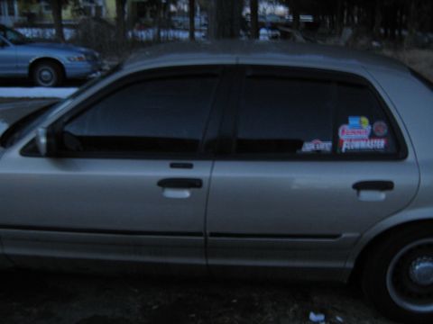 2000 Mercury Grand Marquis - Side view of a silver sedan with tinted windows and aftermarket stickers on the rear passenger window.