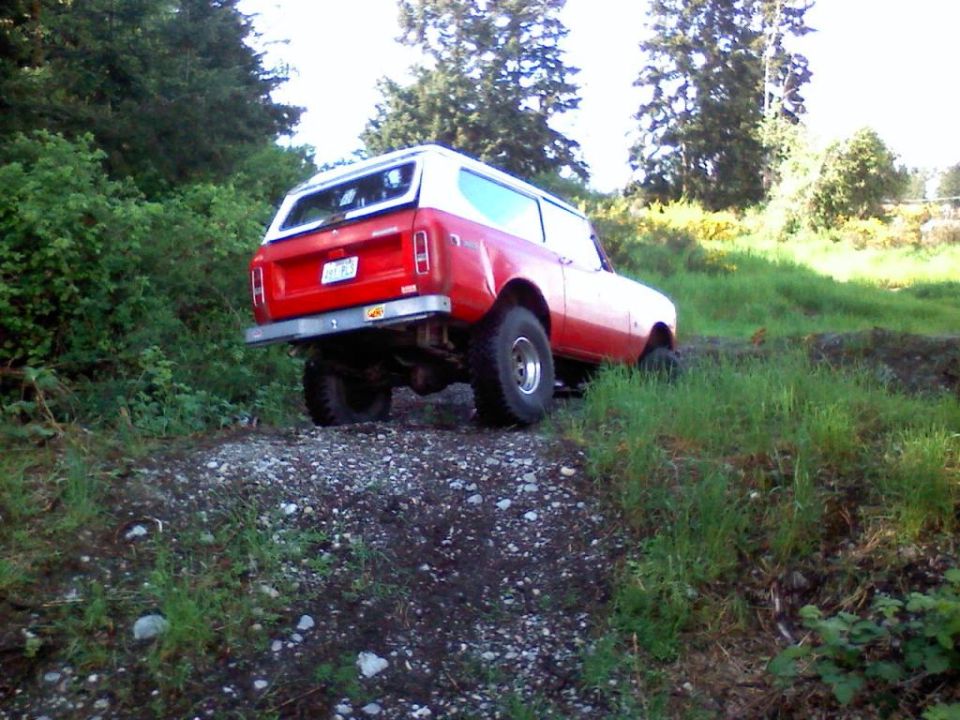Red and white lifted Scout II SUV on a gravel trail in the woods, rear three-quarter view
