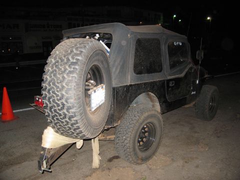 1986 Ford Bronco - A black Jeep Wrangler is viewed from the rear quarter, a soft top and an externally mounted spare tire.
