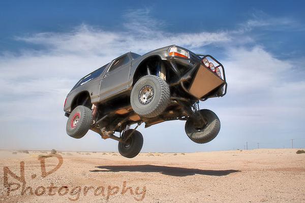 Off-road truck mid-air jump over desert terrain with blue sky and clouds