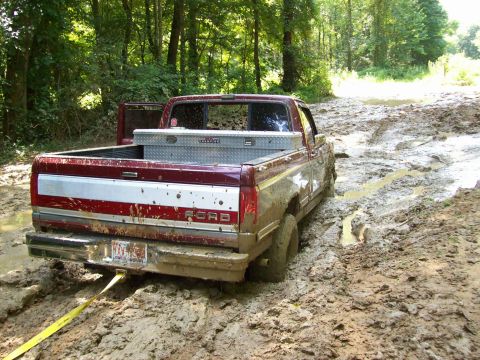 1990 Ford F-150 - A maroon 1990 Ford F-150 is stuck in deep mud on a trail, with a yellow tow strap attached to the rear bumper.
