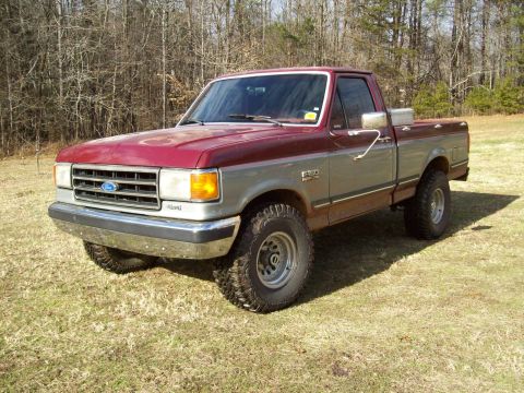 1990 Ford F-150 - This two-tone Ford F-150 pickup truck is shown from the front three-quarter view, parked on a grassy area with trees in the background.