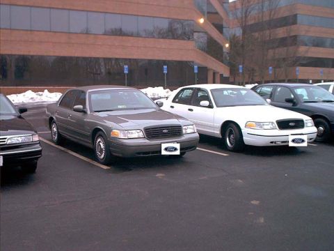 2000 Ford Crown Victoria - Two Ford Crown Victorias are parked in a lot in front of a brick building.