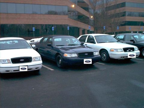 2000 Ford Crown Victoria - Multiple Ford Crown Victoria police cars are parked in a lot in front of an office building.
