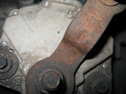 1992 Ford Bronco - Close-up of a rusty steel bracket or arm bolted to a light-colored cast aluminum housing, secured by a large hex bolt with a rounded head.…