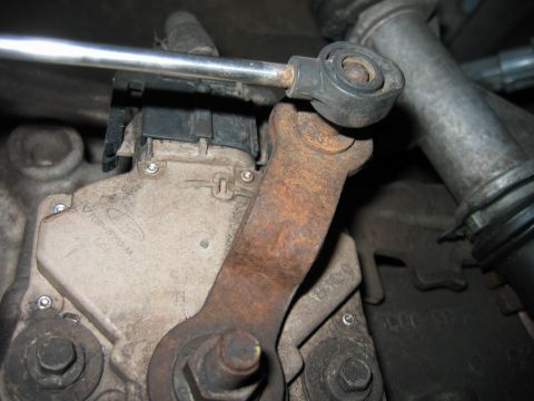 1992 Ford Bronco - Close-up of a rusty metal linkage arm connected to a ball-joint rod end, mounted on a cast component with a Ford logo and part markings. A…