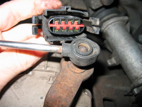 1992 Ford Bronco - A hand holds a black two-piece electrical connector with a red locking wedge and four visible metal terminals, unplugged near a greasy, rus…