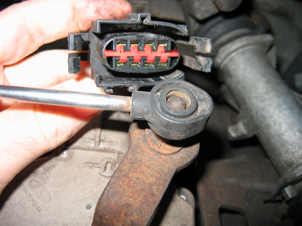 Hand holding disconnected black electrical connector with red lock over an engine bay part