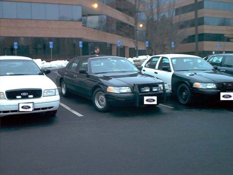2000 Ford Crown Victoria - Several Ford Crown Victoria police cruisers are parked in a lot outside a modern office building.