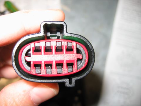 1992 Ford Bronco - Close-up of a hand holding an 8-pin electrical connector with a black outer housing and a red inner terminal grid, viewed head-on at the ma…