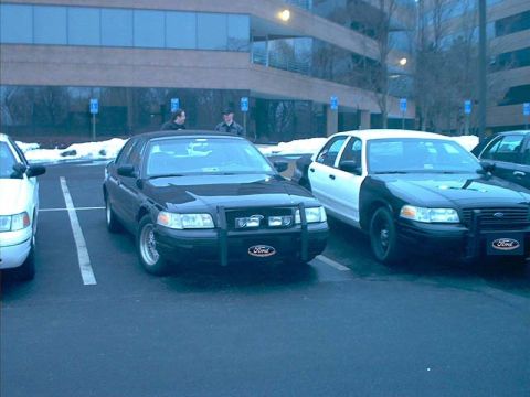2000 Ford Crown Victoria - Two Ford Crown Victoria police cars are parked in a lot, one black and one with a black and white paint scheme.