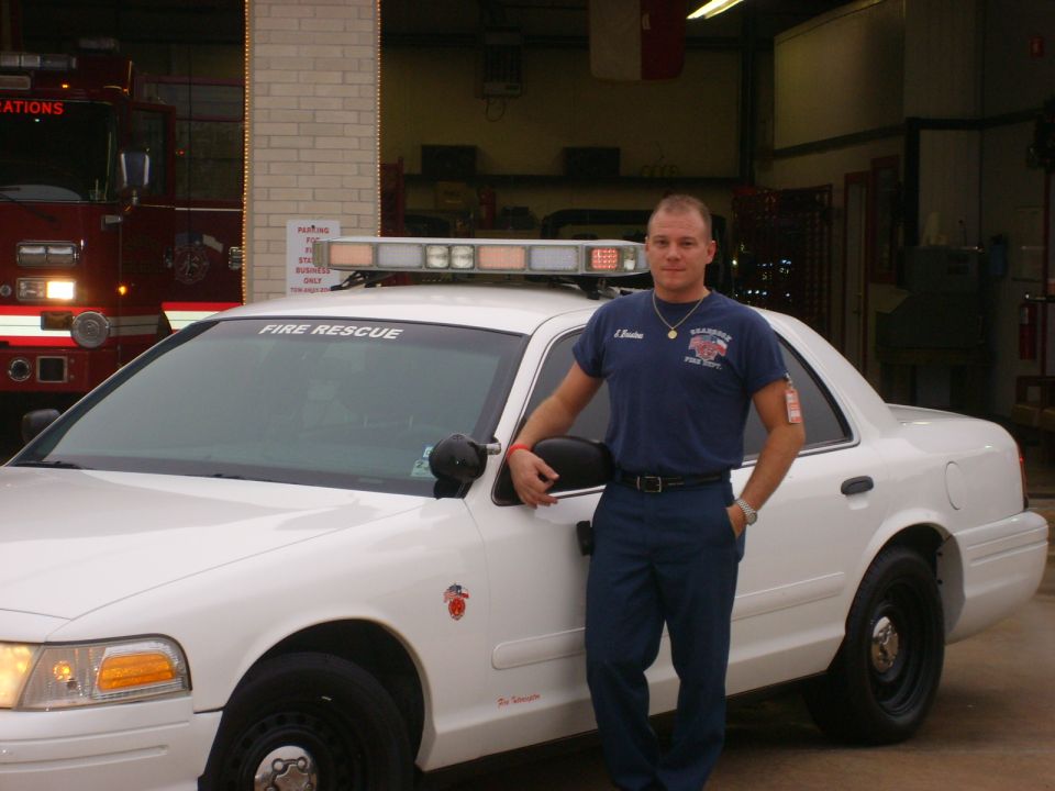 Firefighter stands next to white fire rescue car. Red fire truck in background