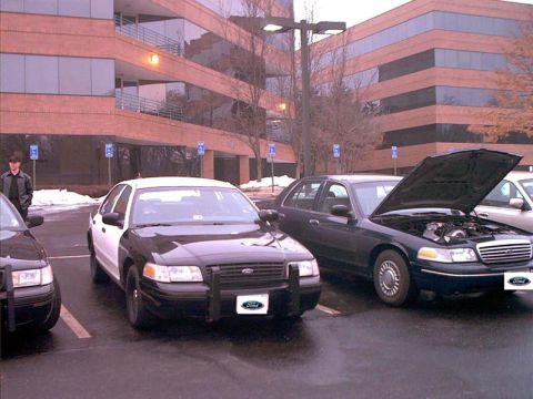 2000 Ford Crown Victoria - Two Ford Crown Victorias are parked in a lot outside a brick building; one is a police car.