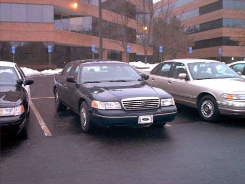 2000 Ford Crown Victoria - A dark blue Ford Crown Victoria is parked in a lot next to a tan sedan, with an office building in the background.