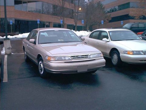 2000 Ford Crown Victoria - A tan Ford Crown Victoria is parked in a lot next to another car, with a modern office building in the background.