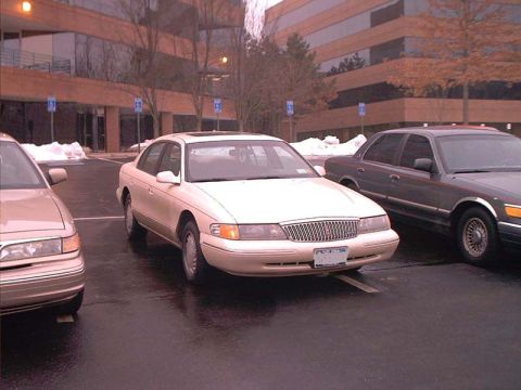 2000 Ford Crown Victoria - A white sedan is parked in a lot next to another vehicle, with a modern office building in the background.